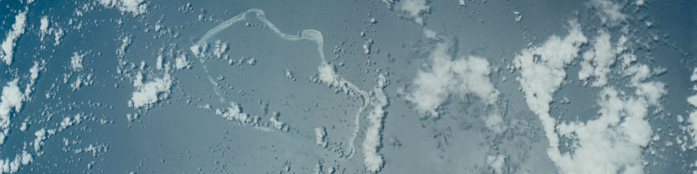 Bikini Atoll from orbit, Gemini 5, August 1965. The rounded bay in the reef at upper left is the crater left by Castle Bravo, detonated 1 March 1954 — two kilometres across, eighty metres deep — here photographed from space for the first time. Original NASA print, S-65-45549.